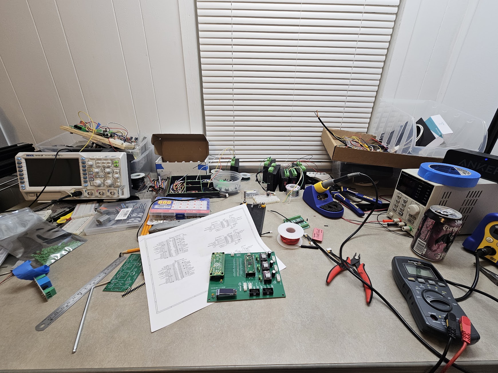 Electronics workbench with oscilloscope, multimeter, soldering tools, printed schematics, and the assembled GardenBot PCB